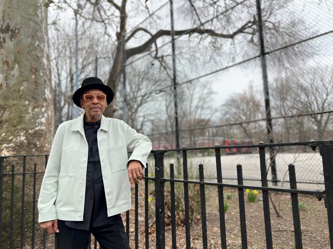 A man in a light jacket, black shirt, and hat stands by a black metal fence in front of a leafless tree and a fenced park area on a cloudy day.