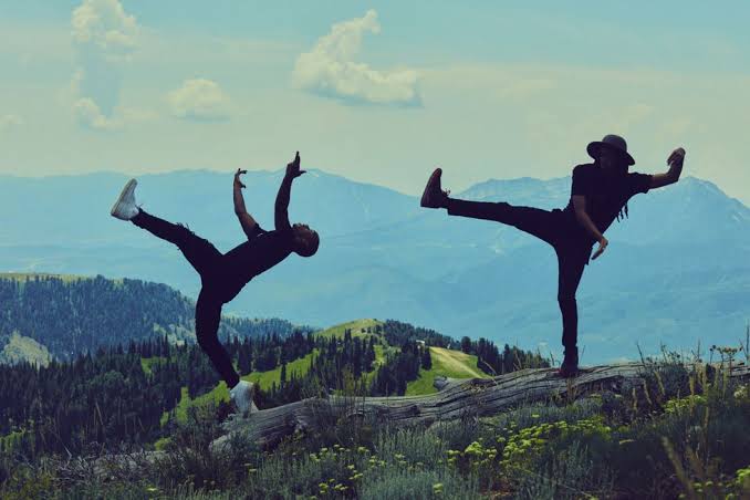 Two people in black clothing balance on a fallen log, striking dynamic, dance-like poses against a scenic mountain background with blue sky and lush greenery.