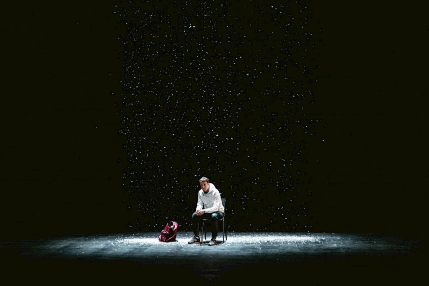 A person sits alone on a chair under falling snow on a dark stage, with a red backpack on the floor beside them, illuminated by a spotlight.