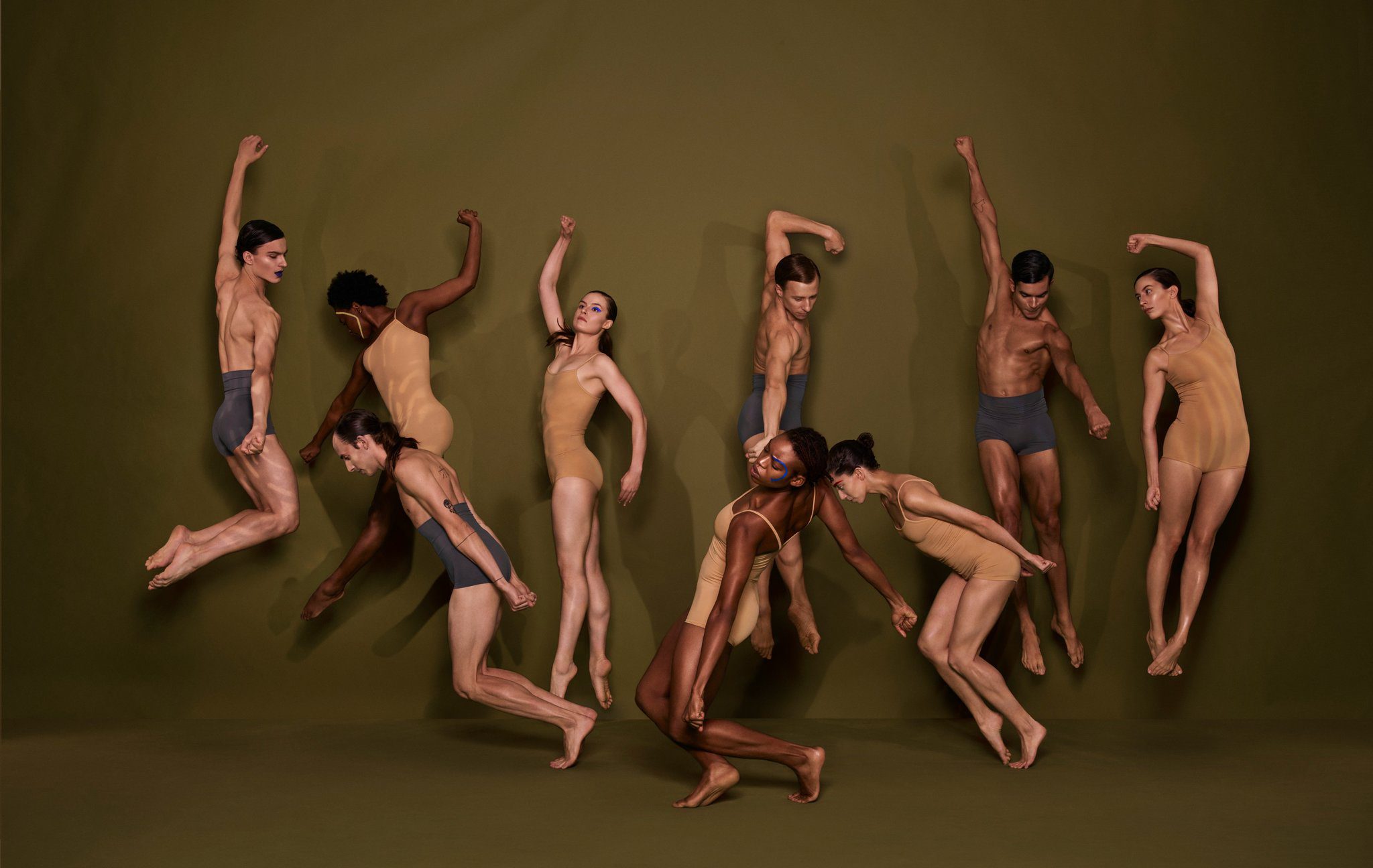Nine dancers wearing tan and gray leotards are captured mid-jump and mid-pose against an olive-green background, creating a dynamic, artistic composition.