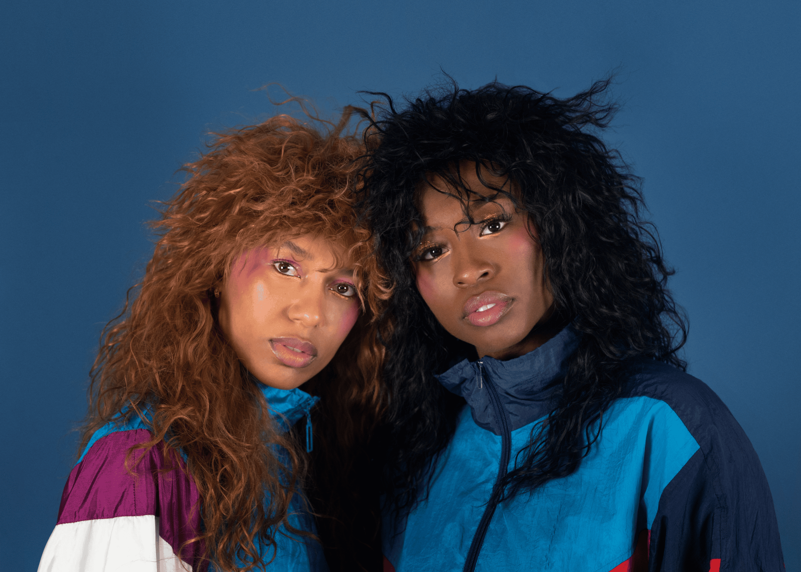 Two women with voluminous, curly 1980s-style hair pose together against a blue background, wearing colorful vintage windbreakers and subtle, vibrant makeup.