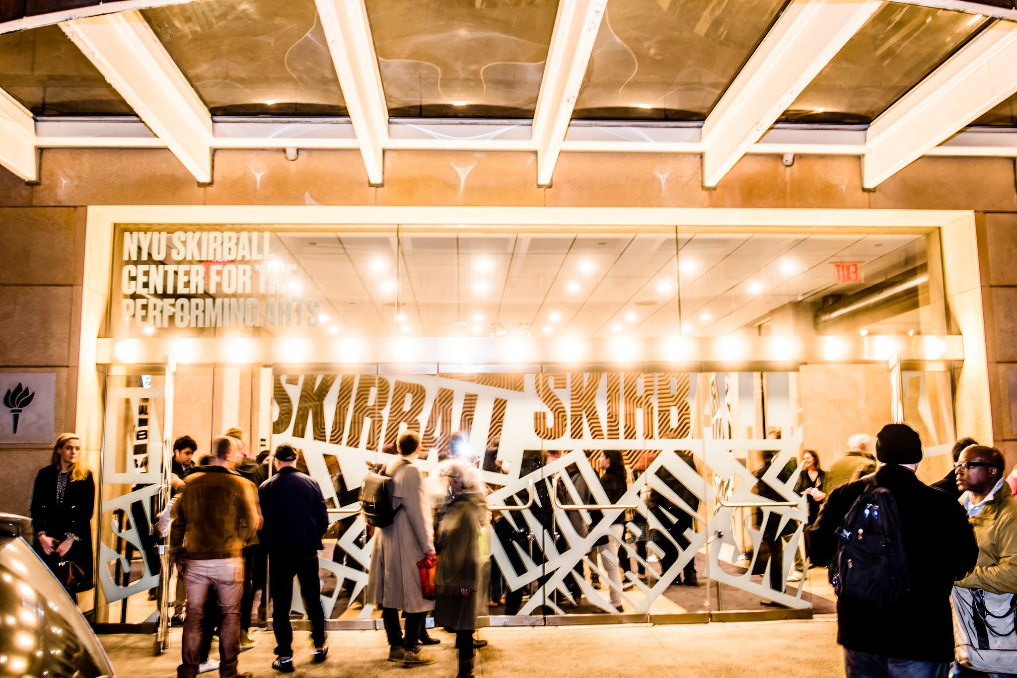 A group of people stand outside the brightly lit entrance of NYU Skirball Center for the Performing Arts at night, with “SKIRBALL” visible on the glass doors.