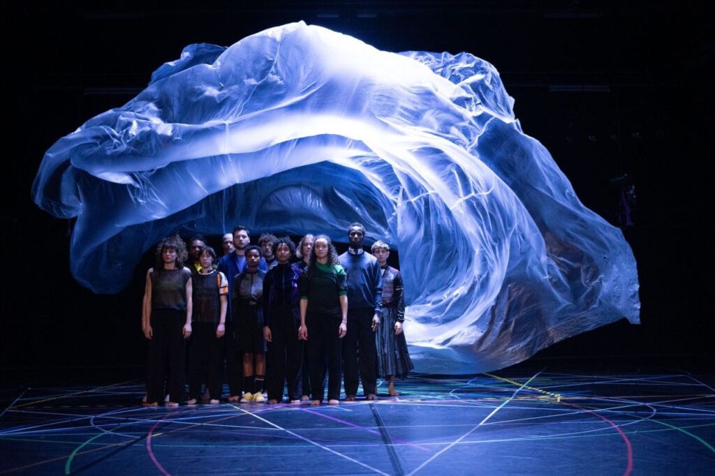 A group of people stands on a stage under a flowing, translucent sheet that is suspended above them, with colored lines on the floor and dramatic lighting creating a surreal atmosphere.