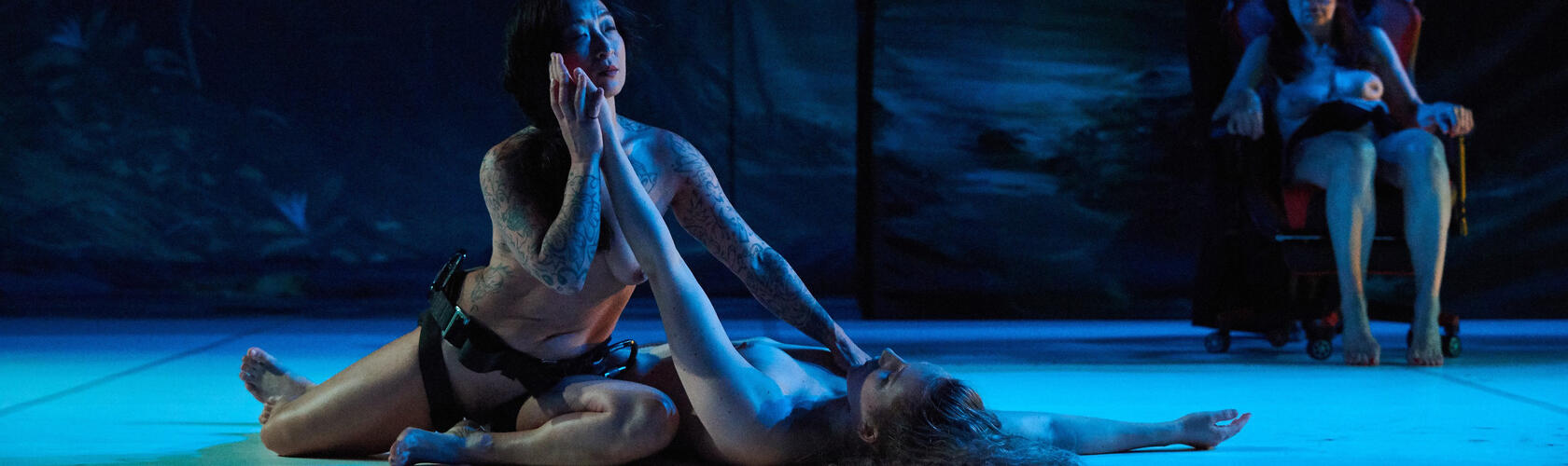 Two performers in dark attire interact expressively on a dimly lit stage, one lying on the floor while the other kneels beside them. A third person sits in a wheelchair in the background under blue lighting.