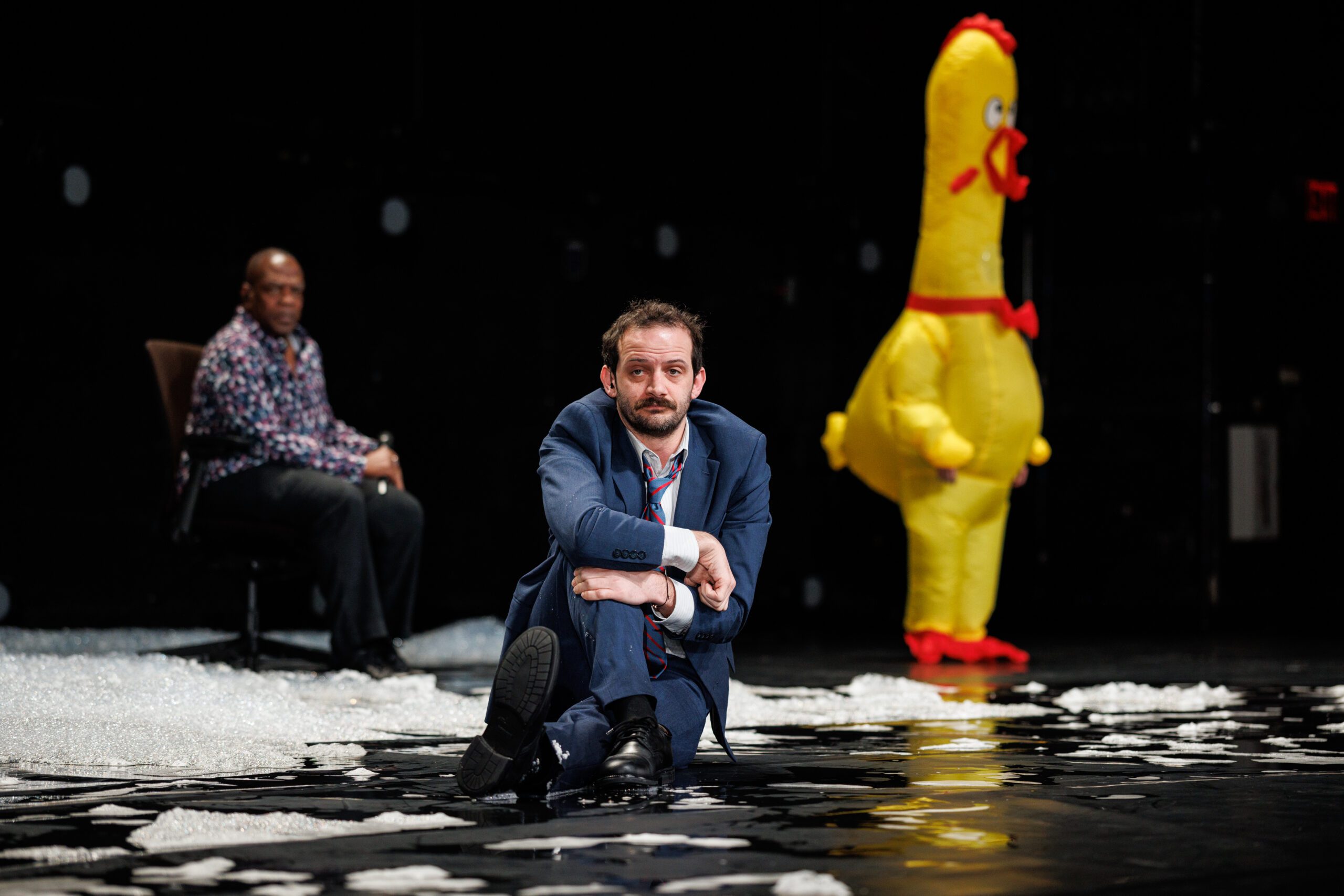 A man in a blue suit sits on the floor with a serious expression, another man sits on a chair in the background, and a person in a giant yellow chicken costume stands behind them on a stage with scattered white material.