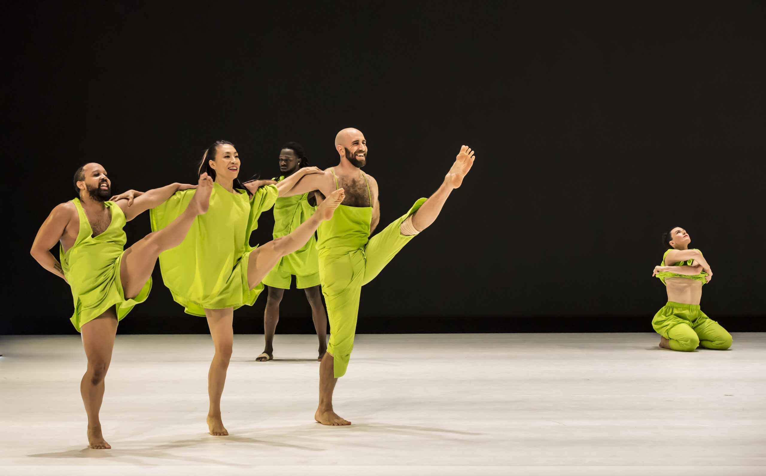 Five dancers in loose, bright green outfits perform on stage. Four are in front, kicking one leg high with joyful expressions, while one dancer is kneeling, arms wrapped around their body, further back and to the right.