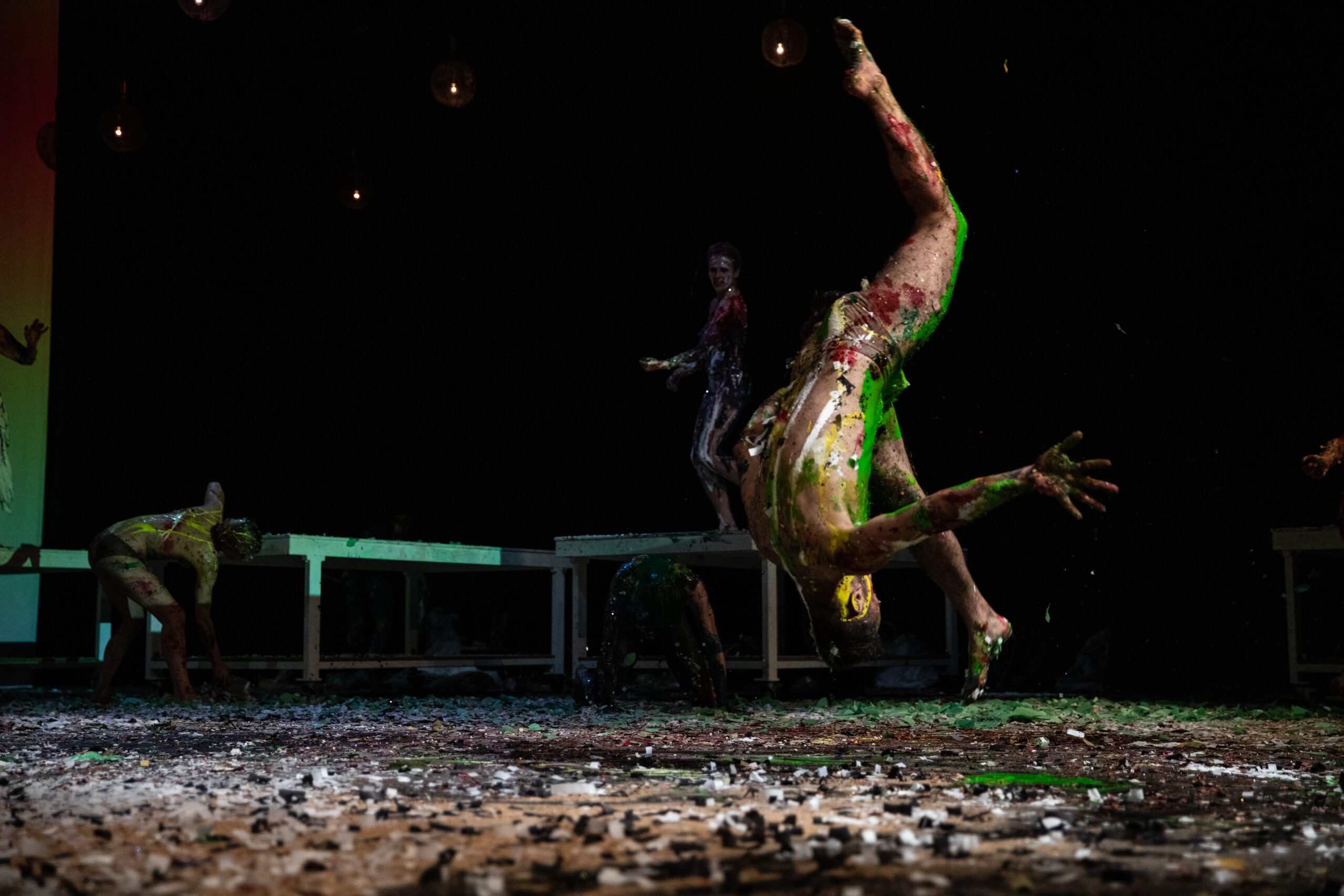 A person covered in colorful paint performs an acrobatic flip on a messy, debris-covered stage, while others move in the background under dim lighting.