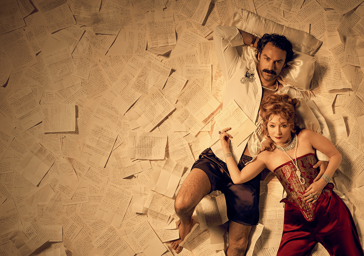 A man and woman lie on a floor covered in handwritten pages. The man wears a white shirt and purple shorts; the woman wears a red corset and holds a pen, resting her arm on the man’s chest. Both look at the camera.