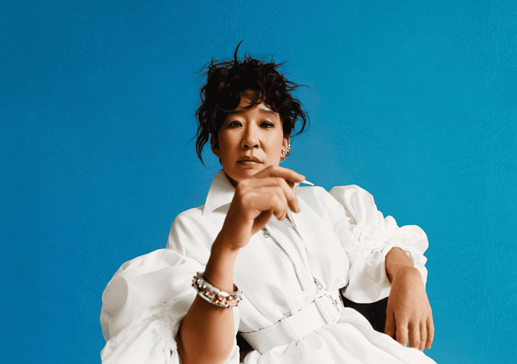Woman with short curly hair sits against a blue background, wearing a white belted dress with puffy sleeves and gold jewelry, looking confidently at the camera.