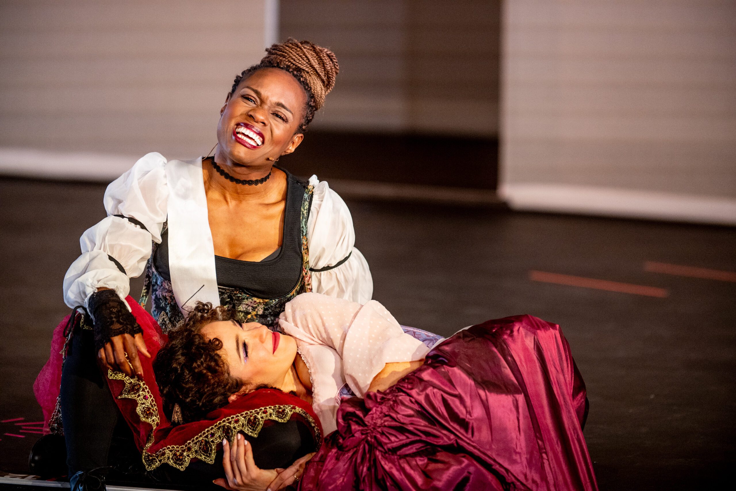 Two women in period costumes perform on stage; one sits upright, smiling widely, while the other lies across her lap in a dramatic pose, wearing a burgundy skirt. The scene is expressive and emotional.