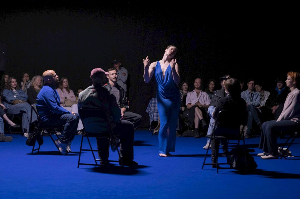 A dancer in a flowing blue dress performs in the center of a seated audience on a blue floor, under dramatic lighting, with people attentively watching.