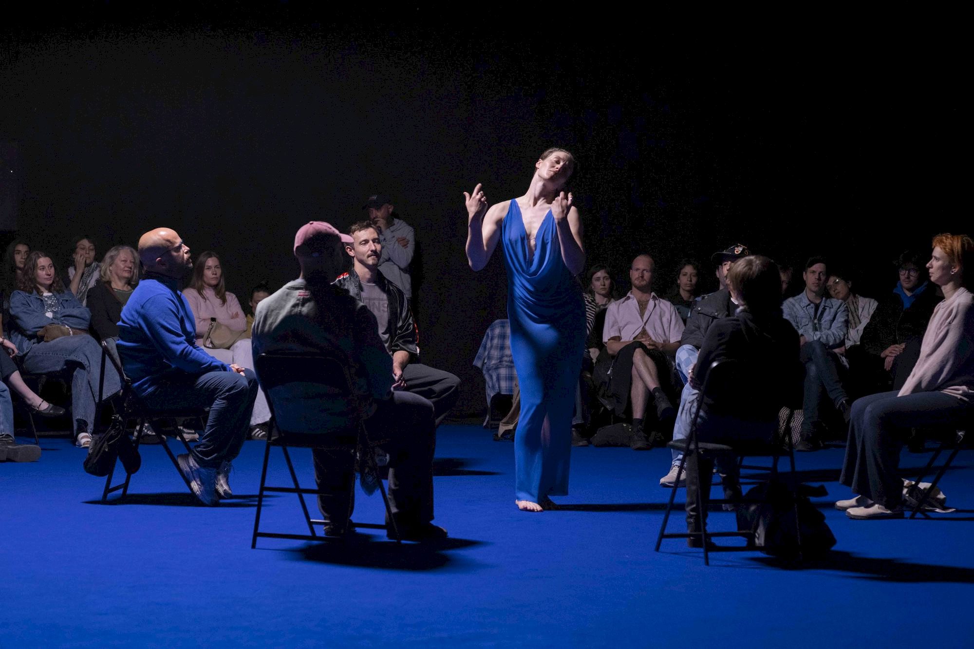 A dancer in a flowing blue dress performs in the center of a seated audience on a blue floor, under dramatic lighting, with people attentively watching.
