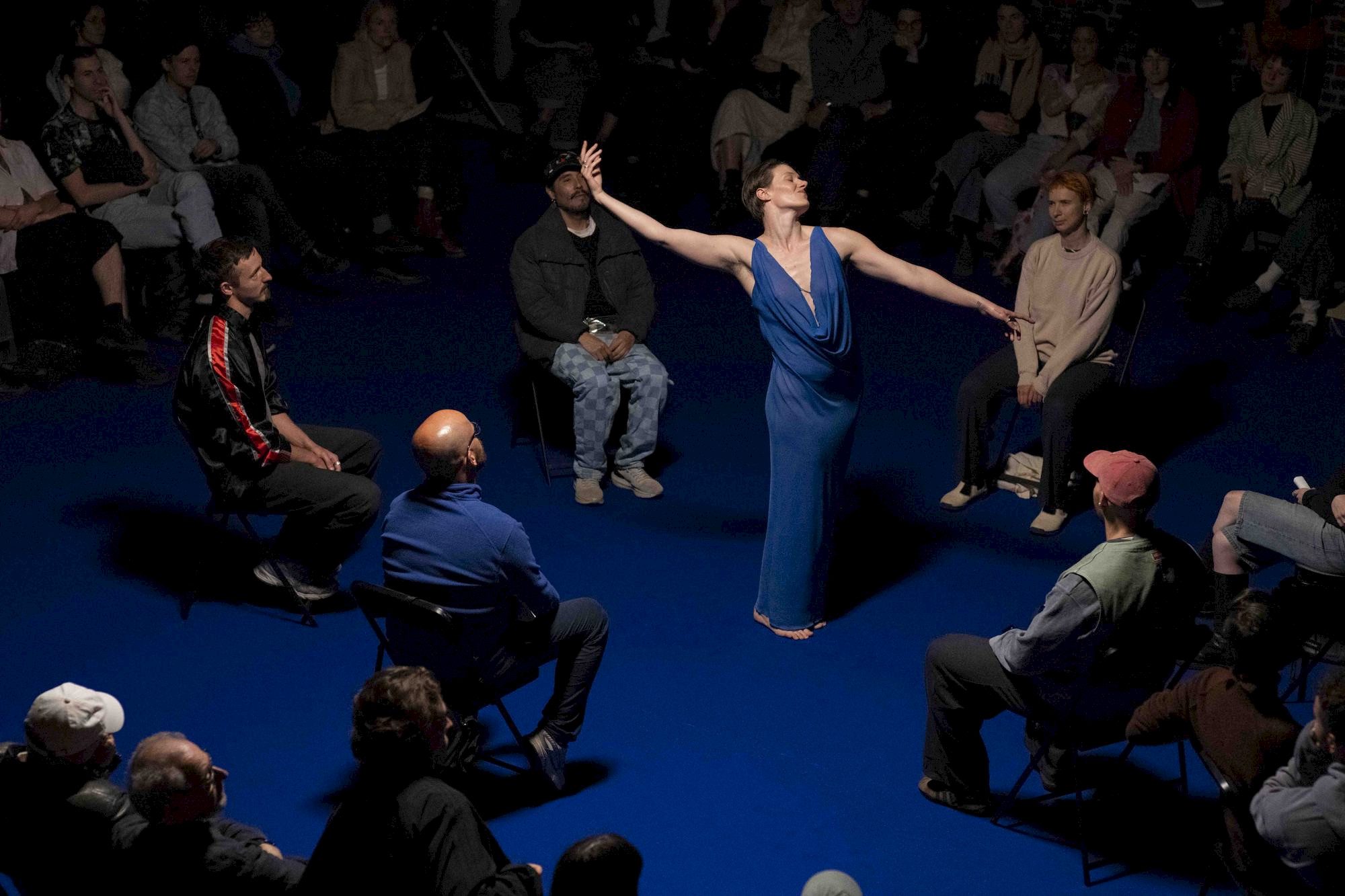 A dancer in a blue dress performs dramatically in the center of a seated audience, arms outstretched, under a spotlight on a blue floor. Spectators watch attentively from all sides.