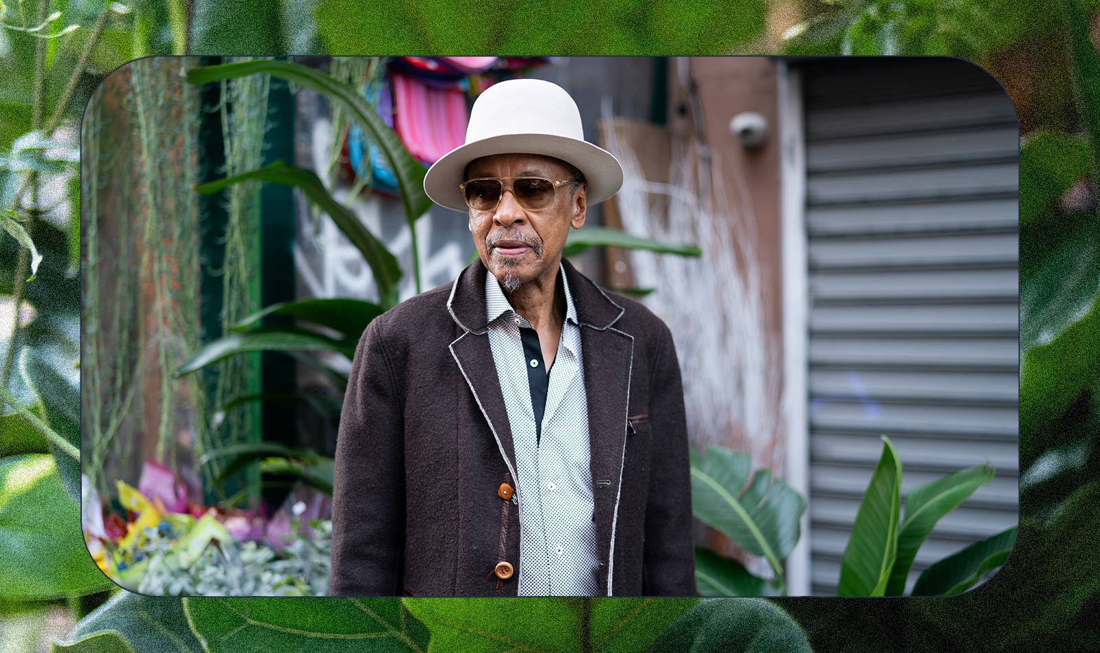An older man wearing a white hat, sunglasses, a dark coat, and a light shirt stands outdoors near plants and colorful decorations, in front of a metal shutter.