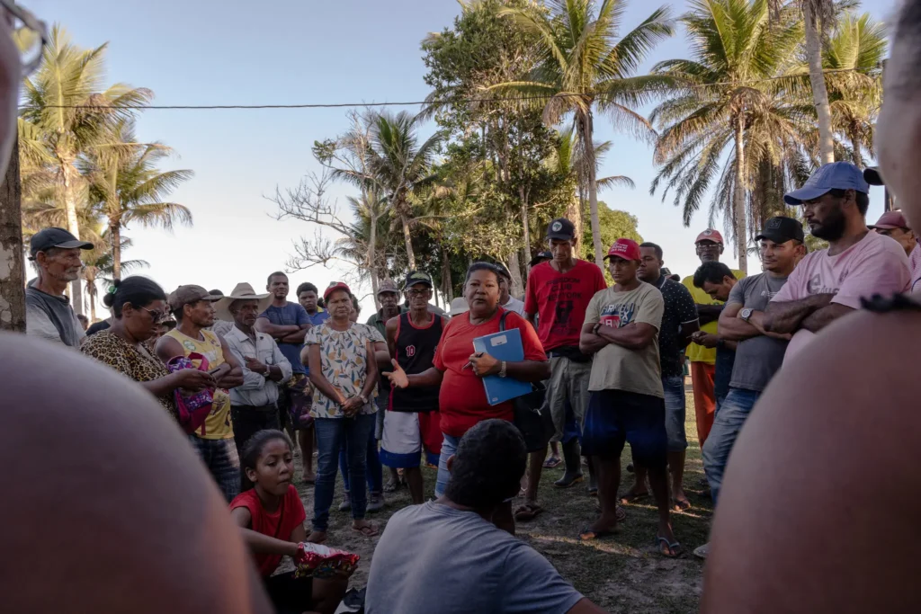 A group of people gathers outdoors near palm trees, listening to a person in a red shirt holding papers at the center. Some stand, others sit, and the mood appears attentive and engaged.