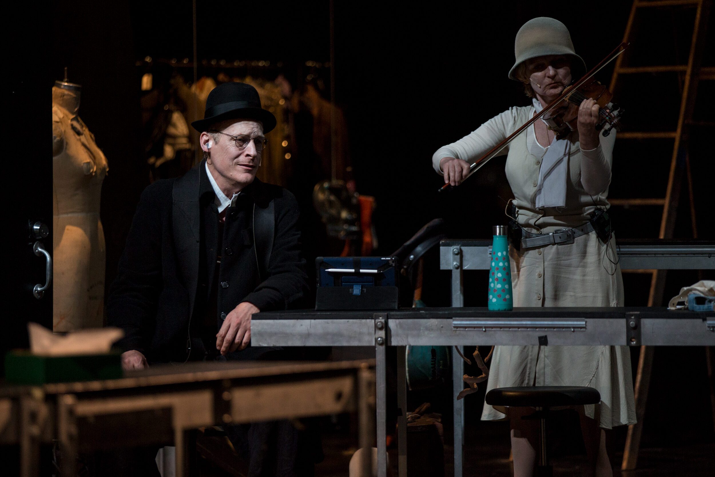 A man in a suit and bowler hat sits at a table, watching a woman in a white dress and hat play the violin. The scene is dimly lit, with mannequins and a ladder in the background.