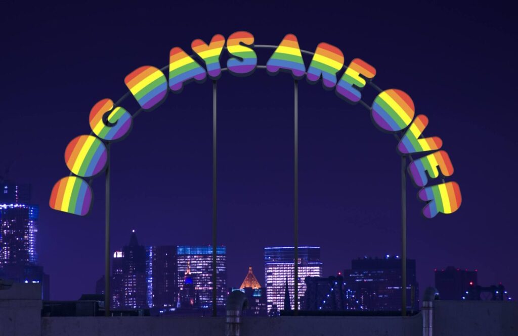 Rainbow-striped 3D letters form an arch reading “DOG DAYS ARE OVER” against a nighttime city skyline with lit buildings.