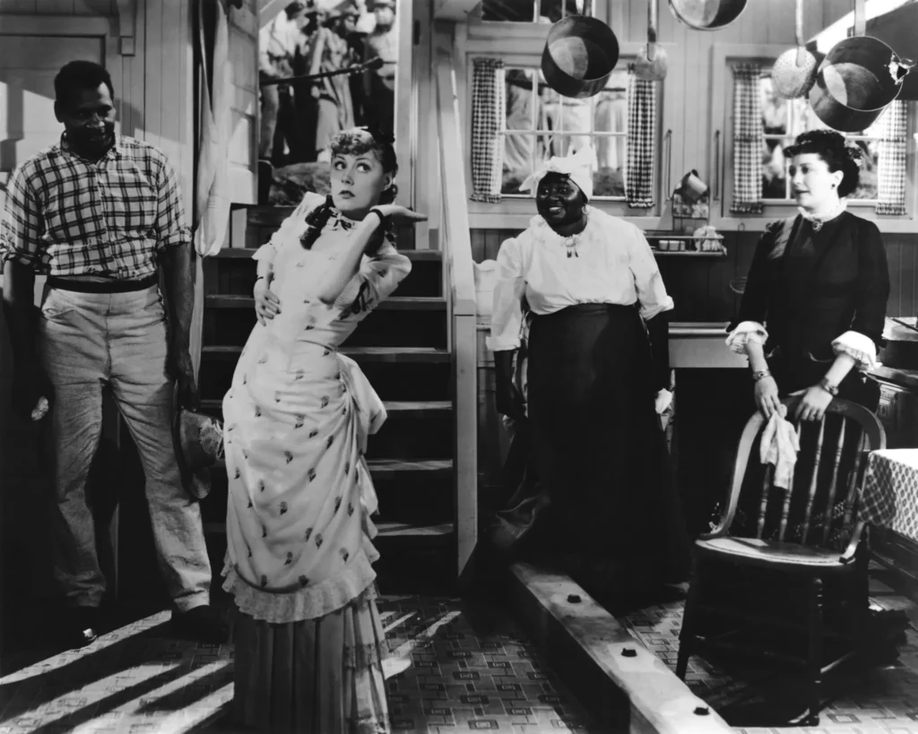 A woman in a white dress dramatically poses in a kitchen with three others watching her; pots hang above, and stairs and a chair are visible.