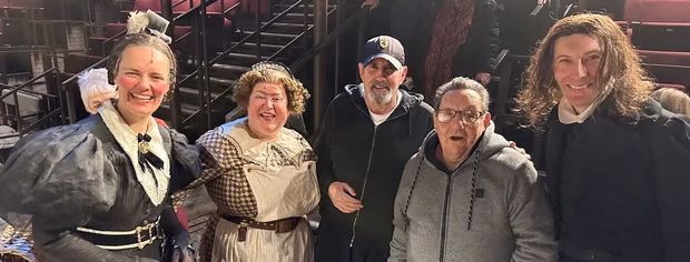 Five people stand together indoors, three wearing historical costumes and two in casual clothes. They are smiling and appear to be enjoying themselves. The background includes theater seats and a staircase.