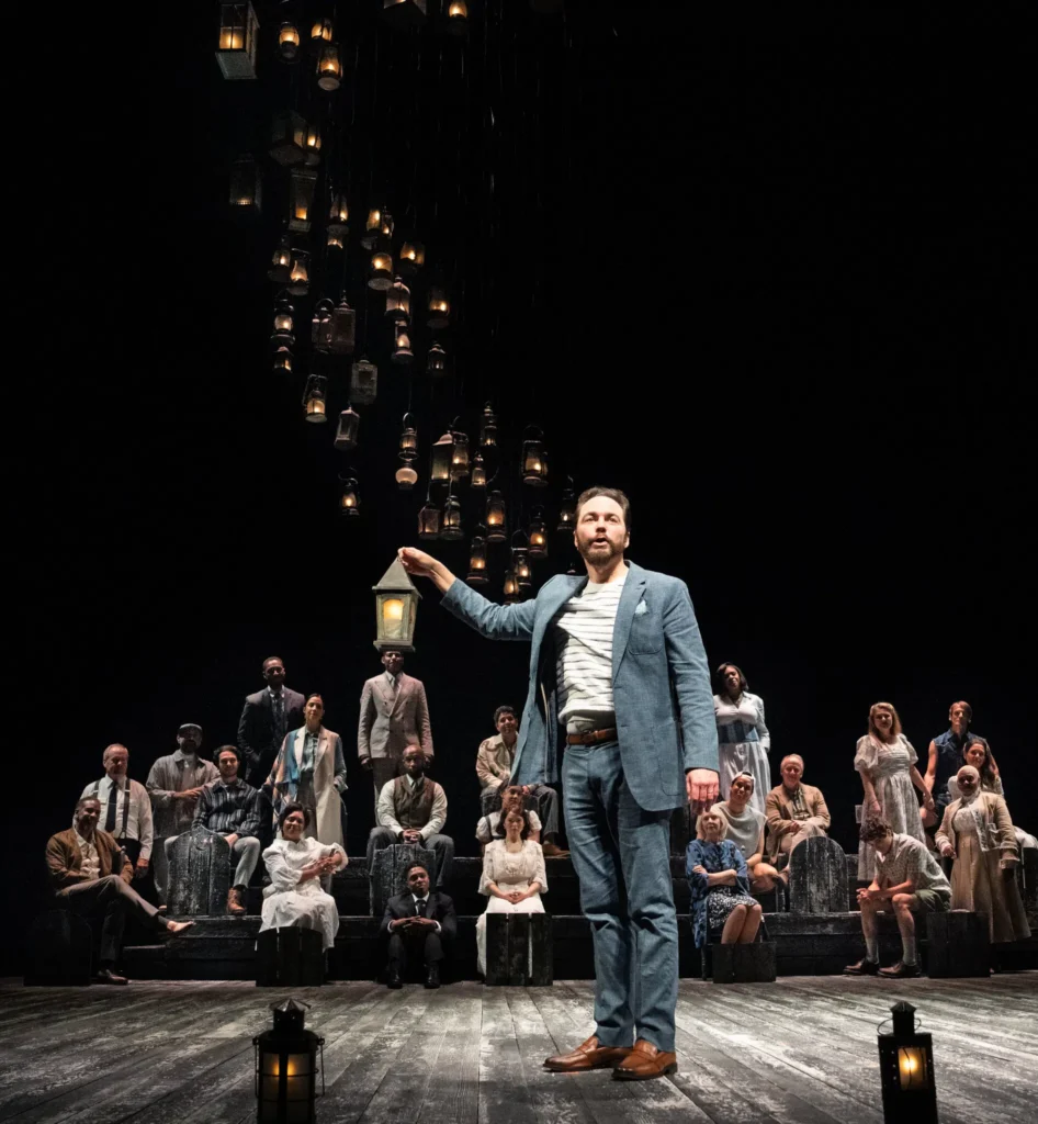 A man in a blue suit stands center stage, arm raised toward hanging lanterns above. Behind him, a group of people in vintage attire sits on tiered benches, watching him. The stage floor is wooden, with lanterns placed in front.