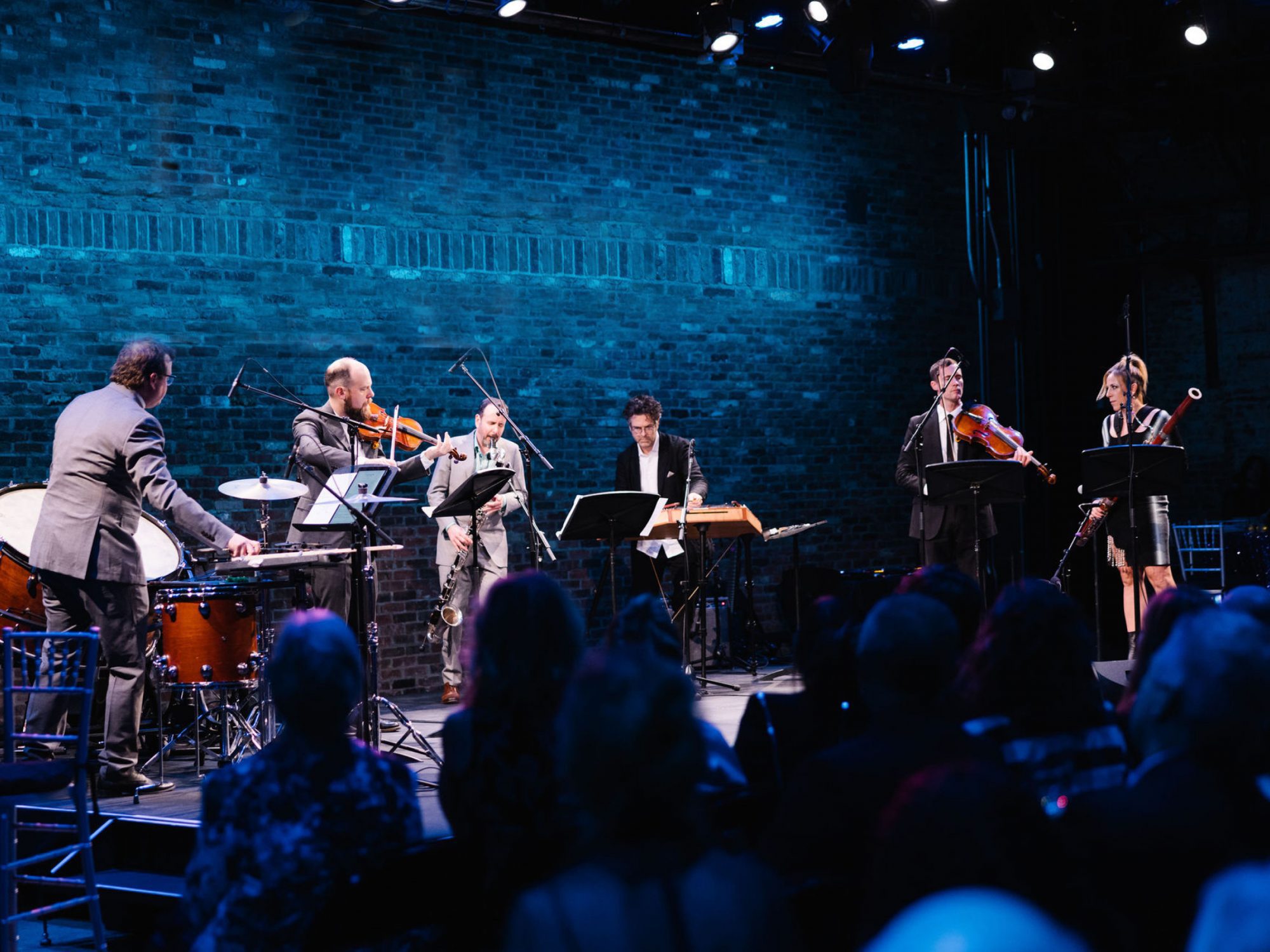 Six musicians perform on stage with various instruments, including drums, violin, and bassoon, in front of a brick wall and seated audience under blue lighting.
