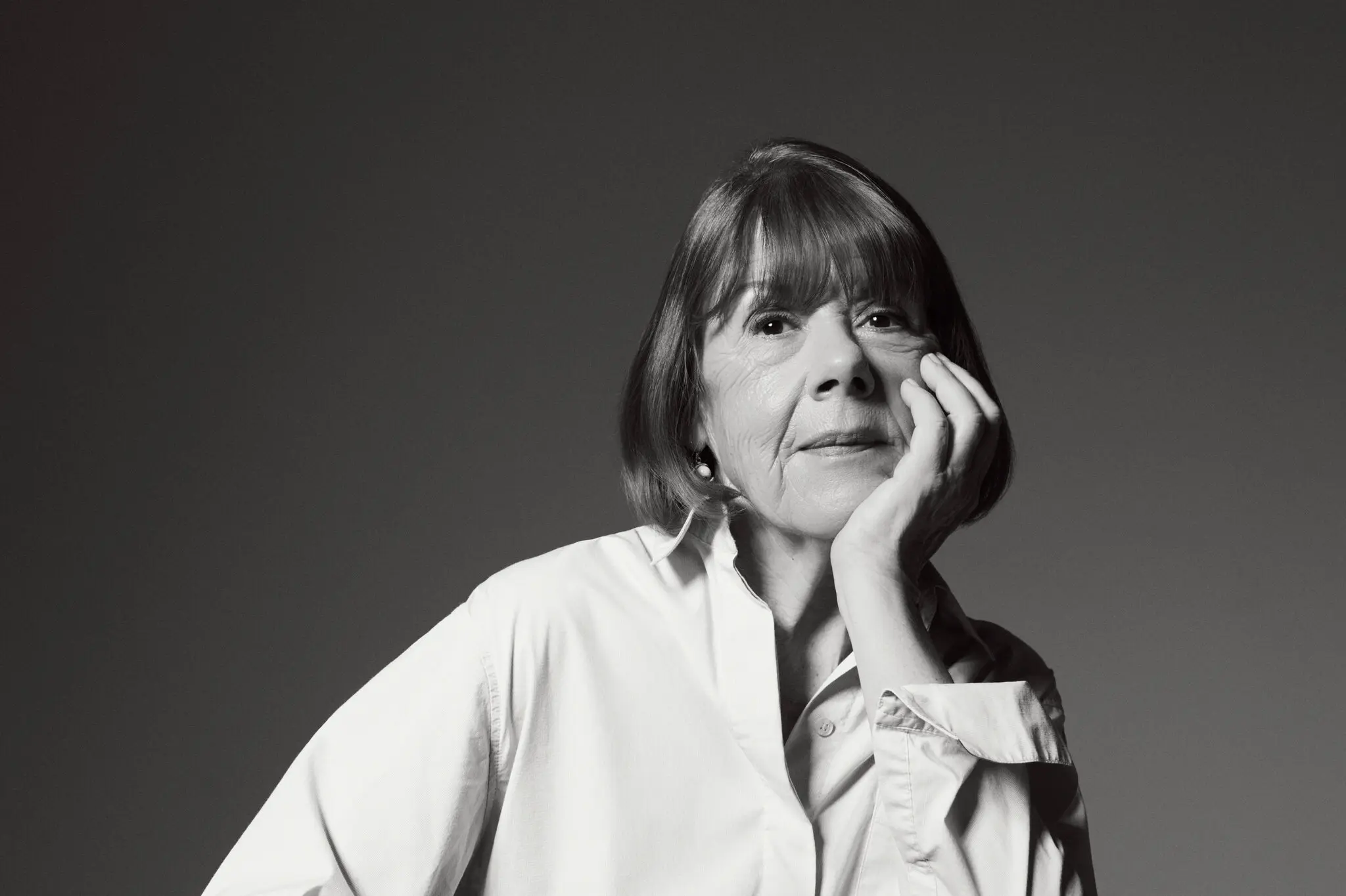 Black and white photo of an older woman with a bob haircut, wearing a light-colored button-up shirt, resting her face on her hand and looking thoughtfully at the camera against a plain background.
