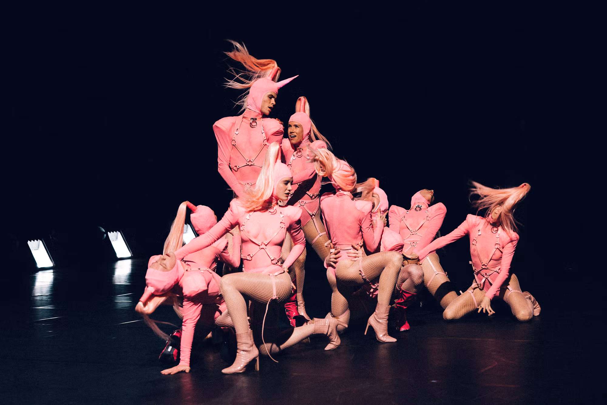 Eight performers in pink costumes and high heels pose dramatically on stage, with some tossing their blond hair. The background is dark, and stage lights shine from the left.