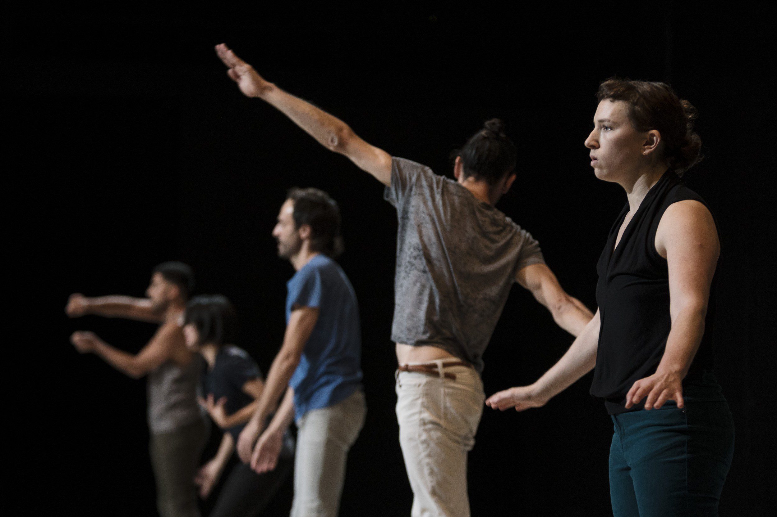 Five people stand in a staggered line, each with one arm extended, appearing to perform a contemporary dance routine against a dark background. Their expressions and gestures vary, suggesting movement and focus.