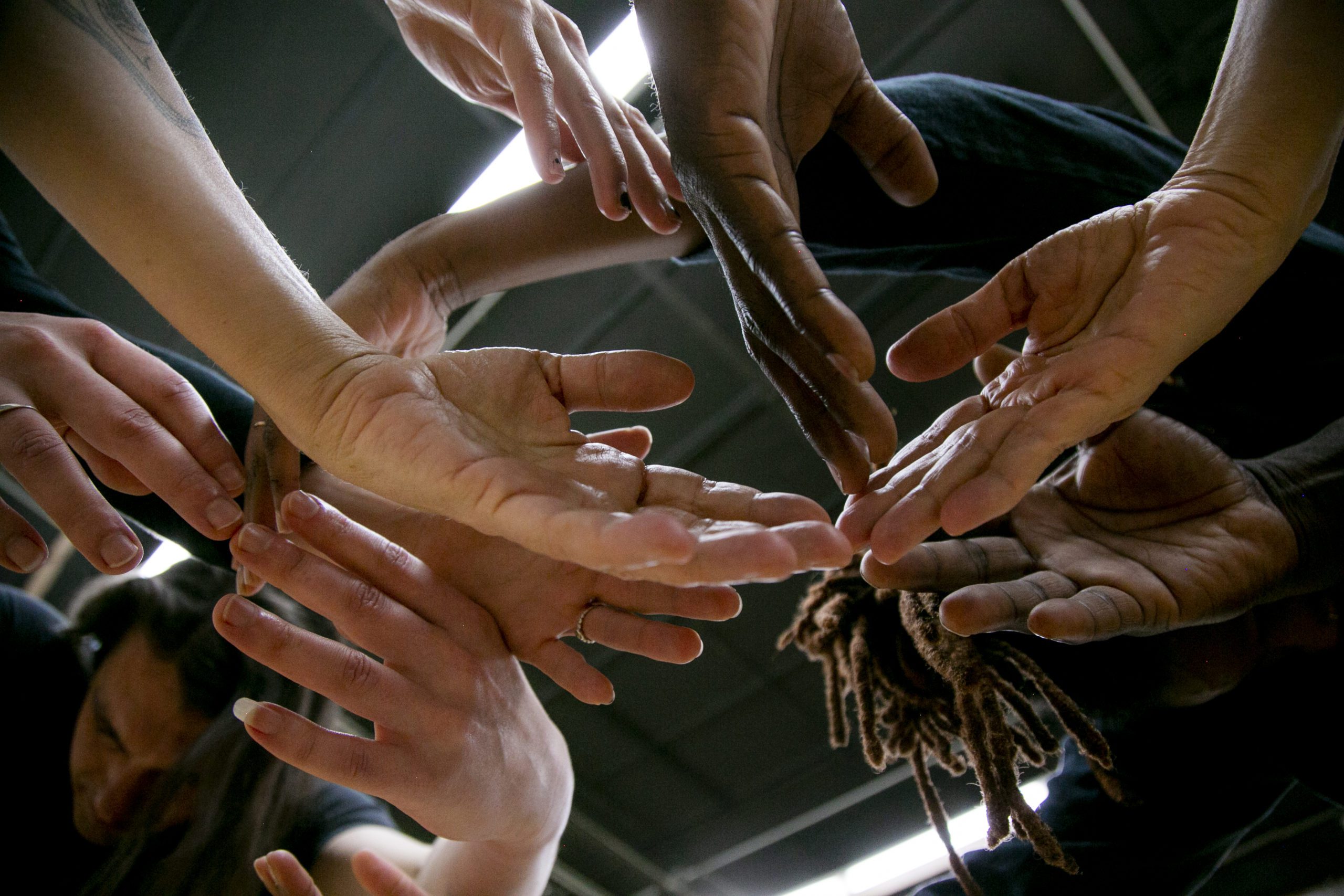 A group of diverse hands reaching toward each other in a circle, viewed from below against an industrial ceiling, symbolizing unity and teamwork.