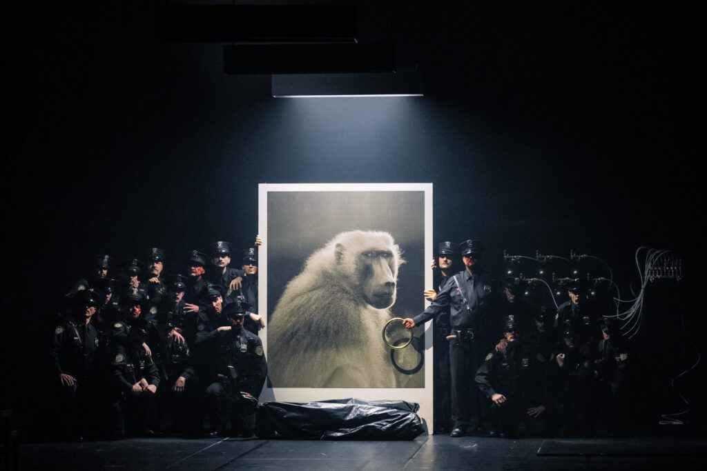 A group of people in dark uniforms pose around a large framed photo of a baboon under a spotlight, with one person holding handcuffs next to the photo.