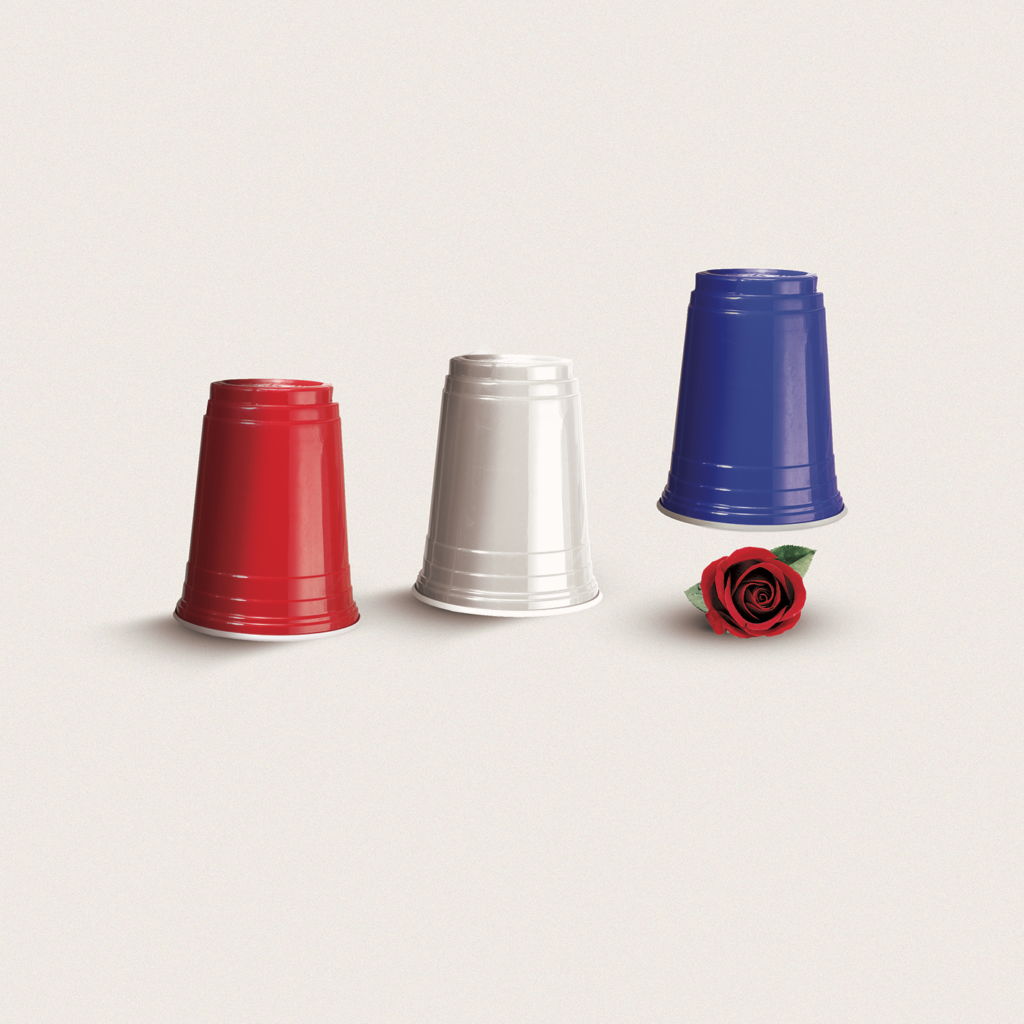 Three upside-down plastic cups—red, white, and blue—stand in a row on a plain background, with a red rose lying next to the blue cup.