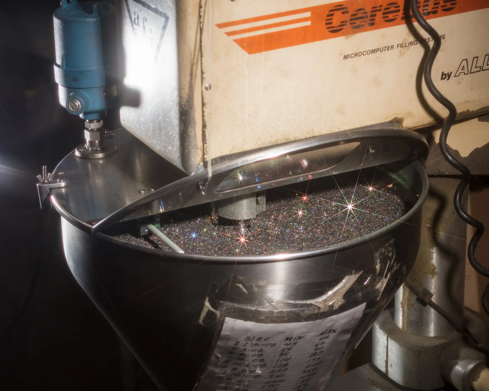 A close-up of a metal industrial machine dispensing shiny glitter-like particles into a large bowl, with light reflecting as small starbursts off the particles.