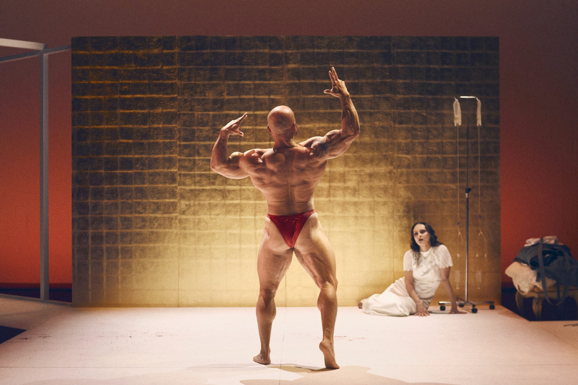 A muscular person in red posing trunks flexes on stage facing a golden wall, while a person in white sits on the floor nearby with an IV stand and bags.