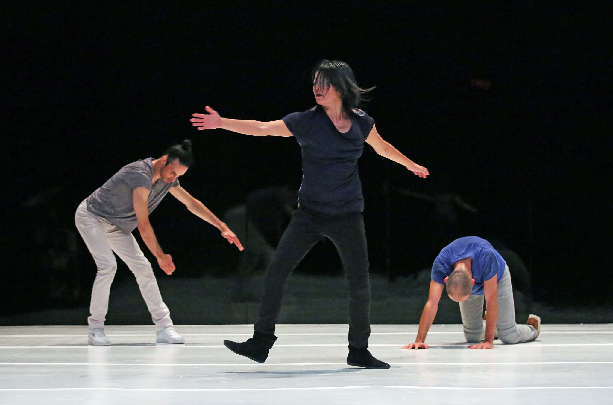 Three people perform a contemporary dance on stage; one stands with arms extended, while the others are bent forward and kneeling, all wearing casual clothes on a white floor.
