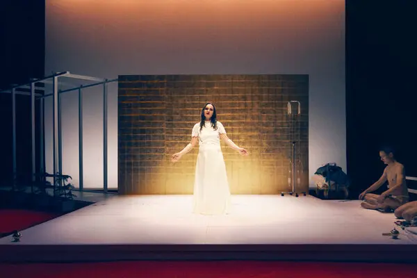 A woman in a long white dress stands center stage on a minimalist set with golden lighting, while a seated person appears to meditate on the right side of the stage.