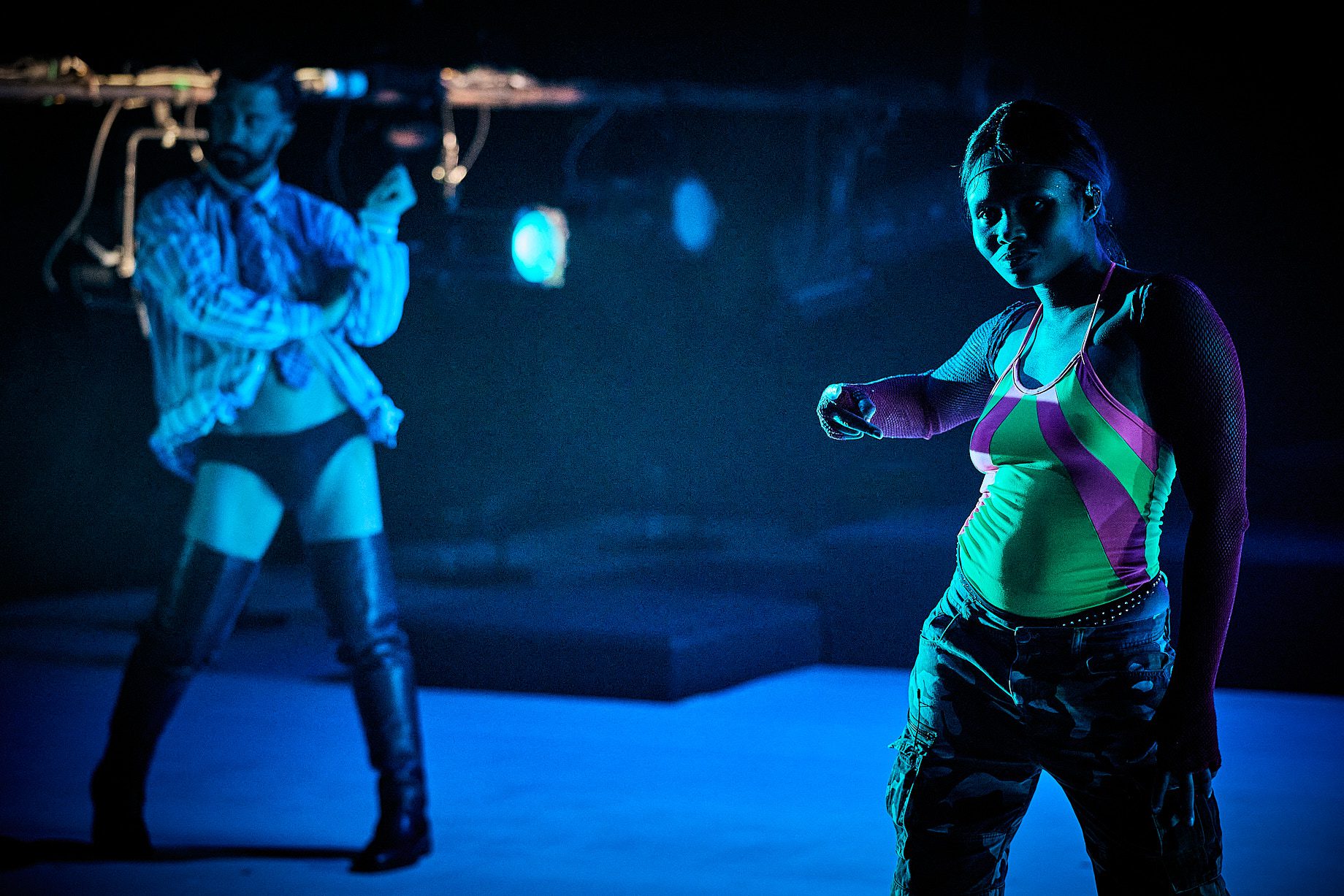 Two performers dance on stage under dramatic blue lighting. One wears a neon bodysuit, fishnet sleeves, and camo pants; the other wears thigh-high boots, shorts, and an open shirt.