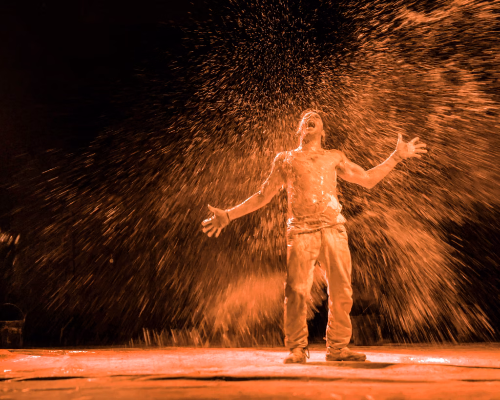 A shirtless person stands center stage, arms outstretched and head tilted back, as powder dramatically bursts around them in orange lighting.