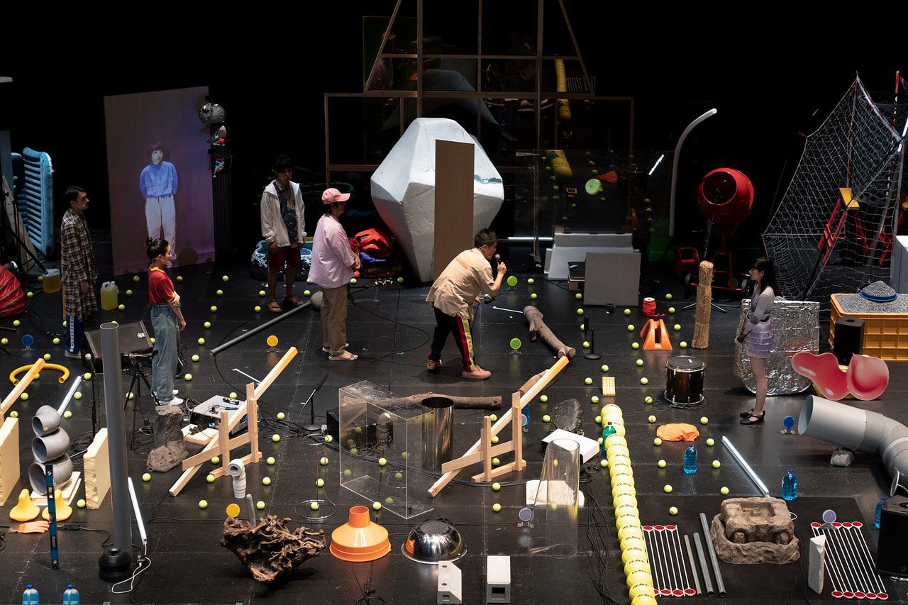 A group of people stand on a stage filled with scattered props, yellow tennis balls, and various objects, creating a chaotic, experimental scene with performance art elements.