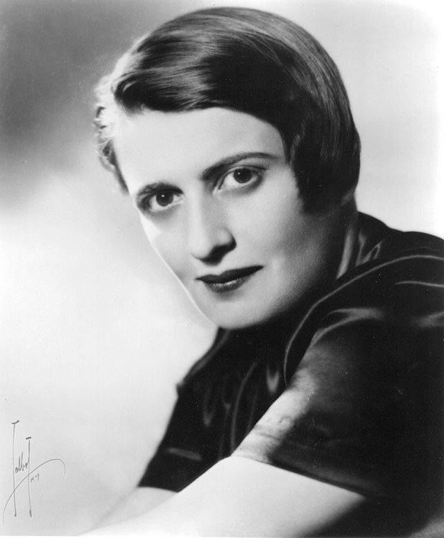 Black-and-white portrait of a woman with short, smooth dark hair, side-parted, wearing a dark, satiny top, looking directly at the camera with a neutral expression.