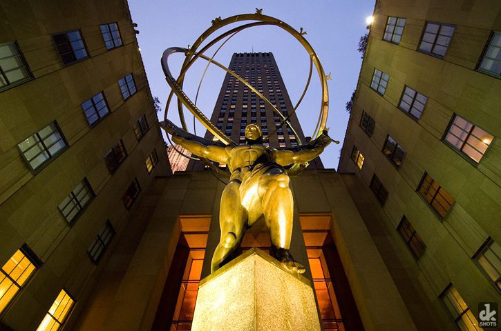 Bronze statue of Atlas holding a large armillary sphere, seen from below at Rockefeller Center in New York City, with a tall building and twilight sky in the background.