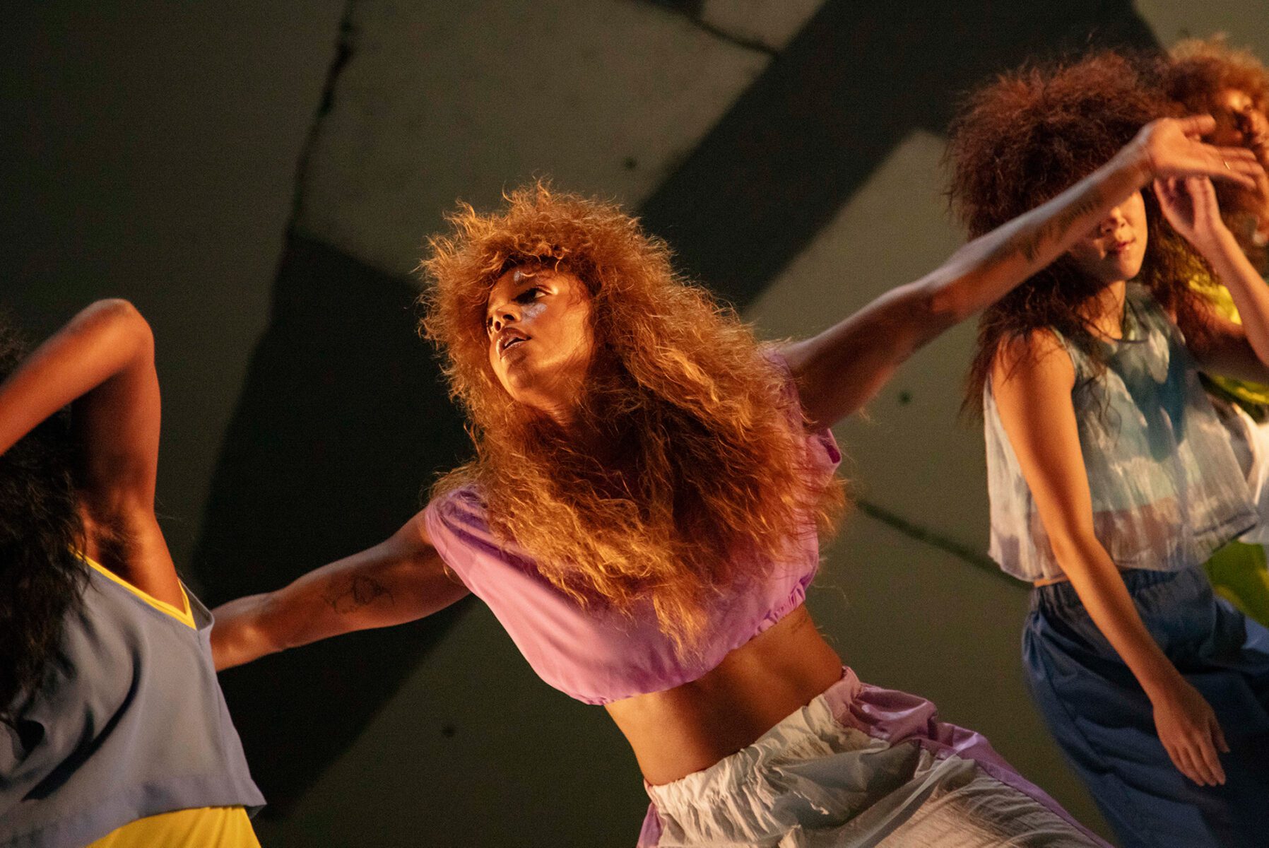 Three dancers with voluminous hair perform expressively on stage, wearing colorful, loose-fitting costumes. The central dancer, in a light purple top, extends her arms, conveying motion and emotion in dramatic lighting.