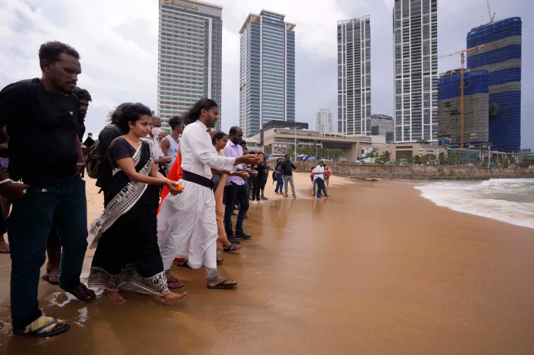 A group of people stand on a sandy beach near tall city buildings, holding items and facing the ocean under a cloudy sky, as if performing a ritual or ceremony.