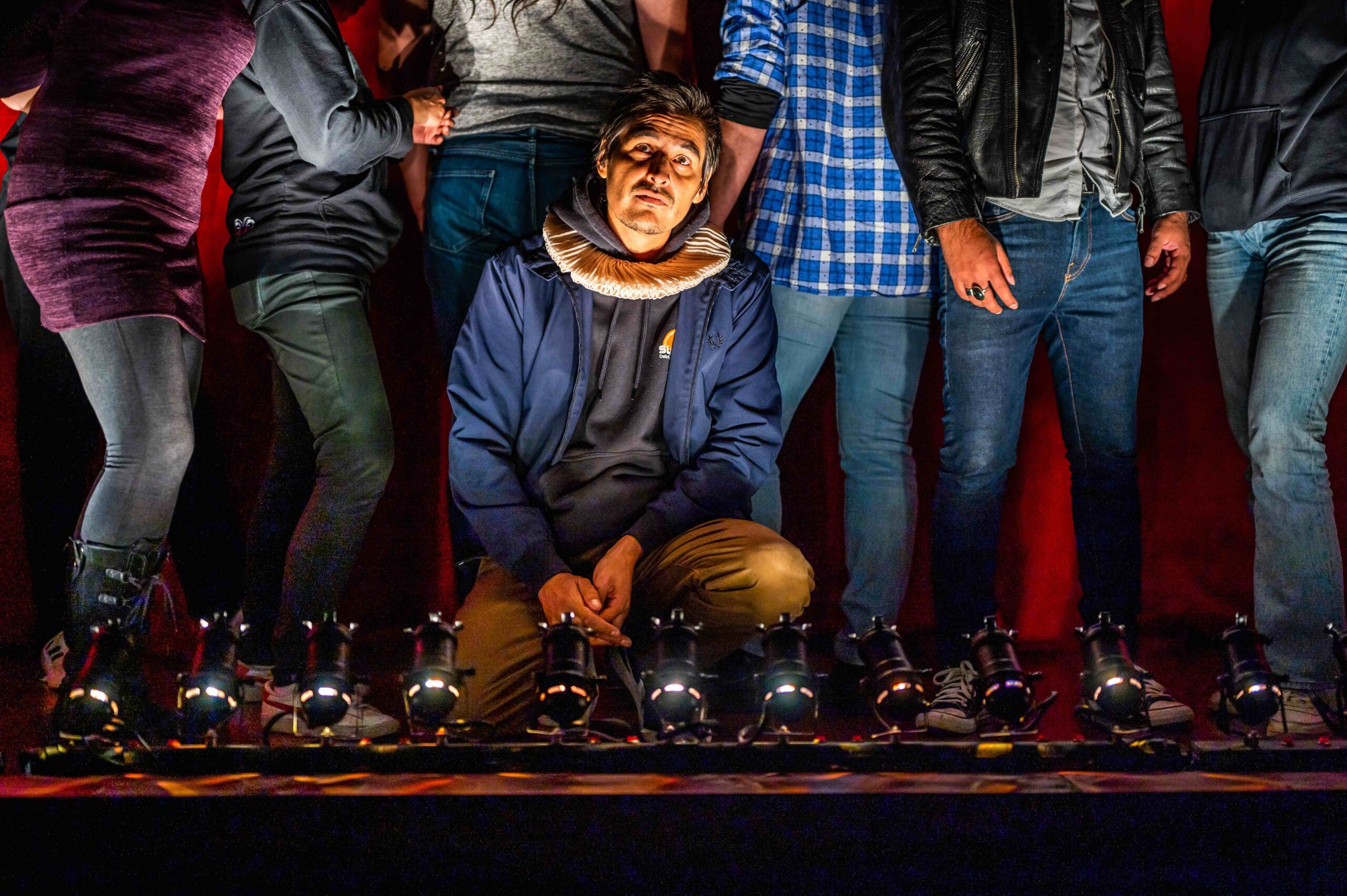 A person wearing a ruff collar crouches under stage lights, surrounded by people in casual clothing. The background is dark red, with dramatic lighting highlighting the central figure’s face.