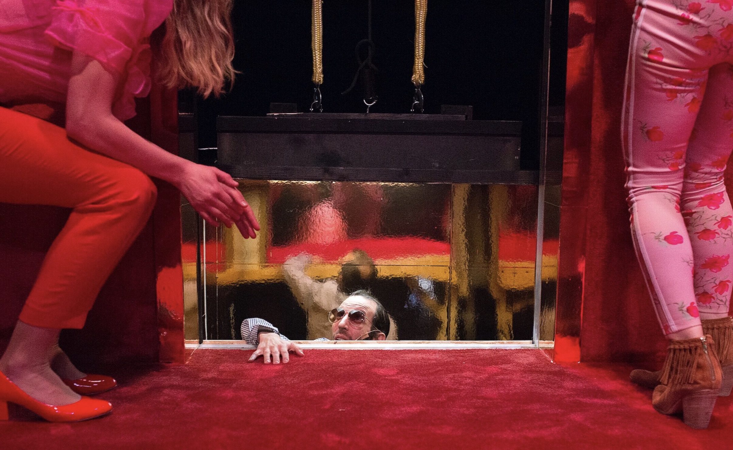 A person in sunglasses peers up from a hole in a red carpeted floor, reaching out while two people in colorful clothing stand on either side, their faces unseen.