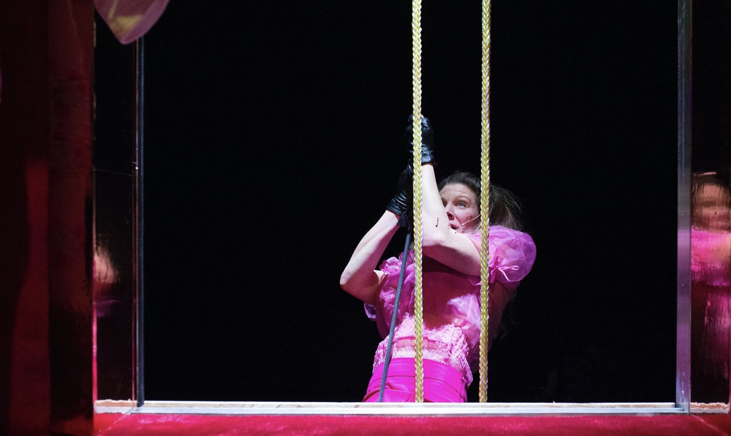 A woman in a bright pink dress and black gloves holds onto two yellow ropes, appearing to be on stage in a theatrical performance, with a dark background and reflections visible on both sides.