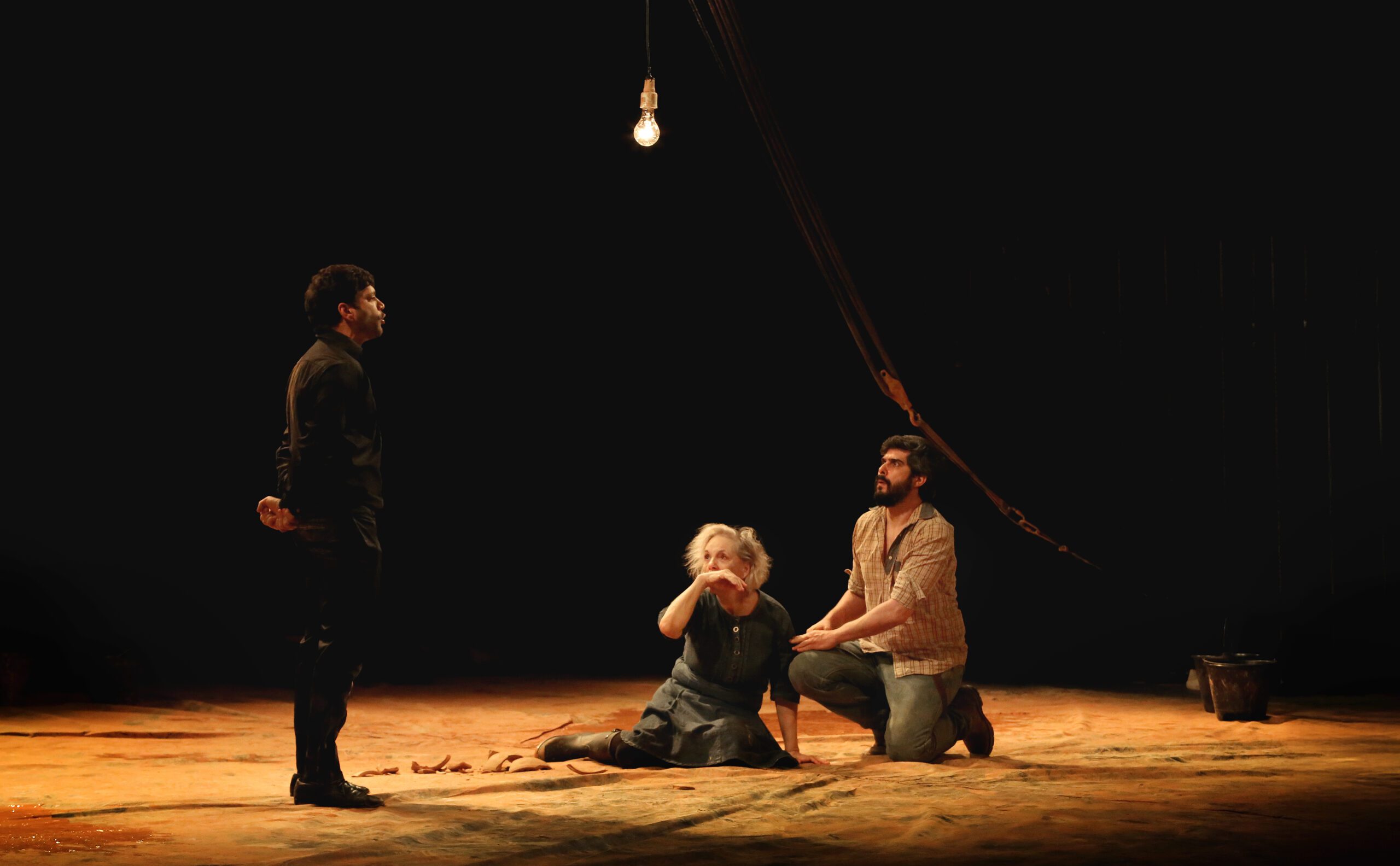 Three actors perform on a dimly lit stage with a single bulb. One stands in black, while two others kneel on the ground, with the woman appearing distressed and the man supporting her. The background is dark.