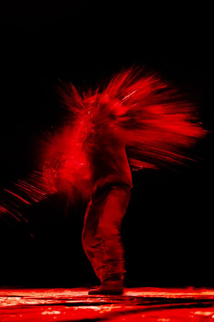 A person stands as a burst of red powder or liquid explodes outward from their body against a black background, creating a dramatic visual effect.