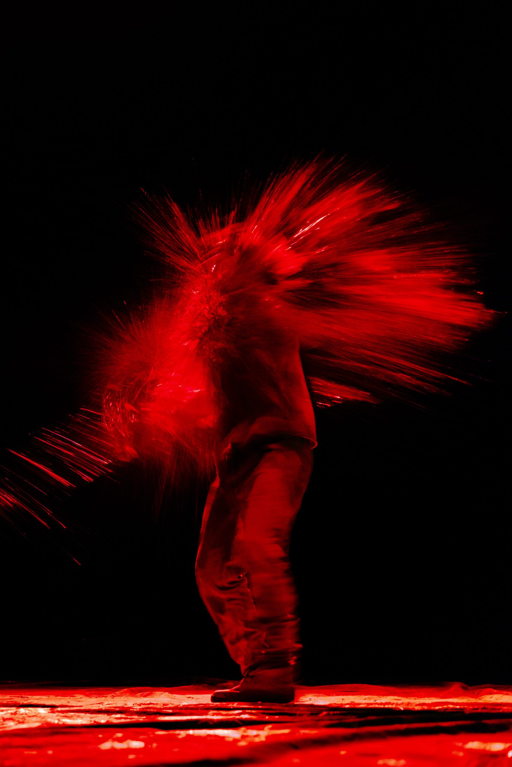 A person stands as a burst of red powder or liquid explodes outward from their body against a black background, creating a dramatic visual effect.