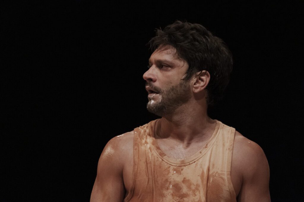 A man wearing a stained tank top, with some white substance on his face and neck, looks to his left against a dark background.