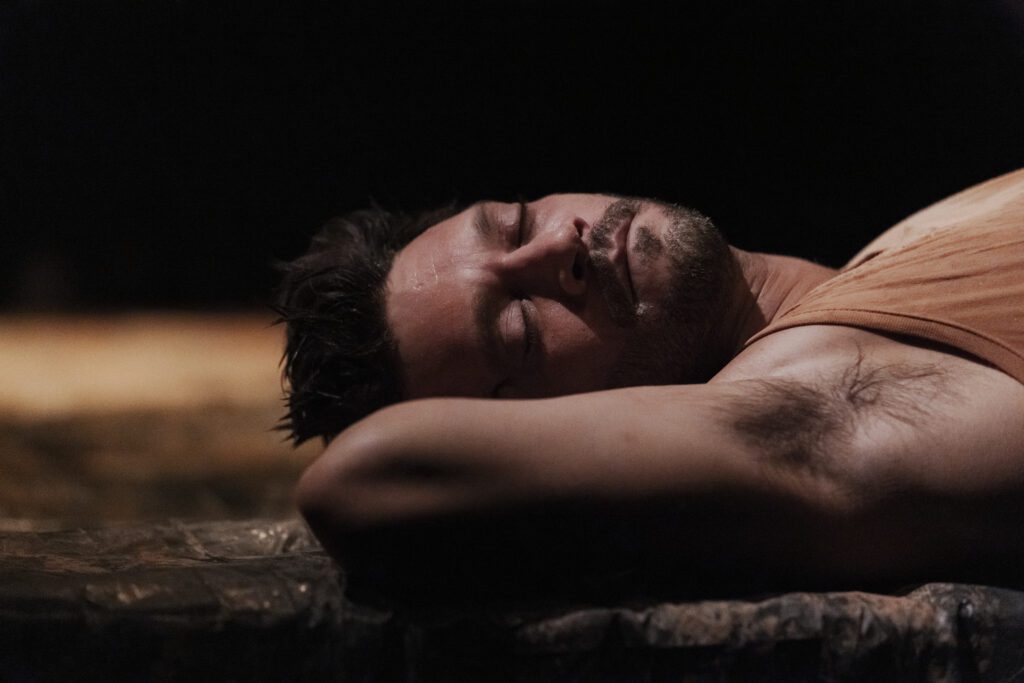 A man with a beard and mustache lies on his side, eyes closed, resting his head on his arm in a dimly lit setting.